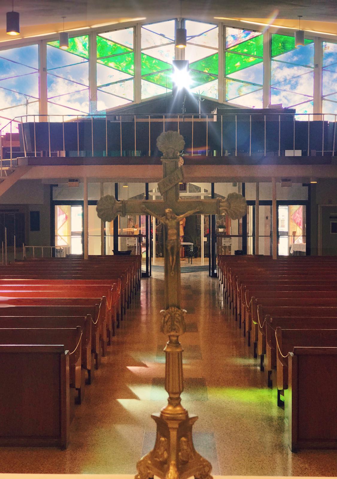 Cross on altar with sun coming through stained glass windows. View of pews in background.